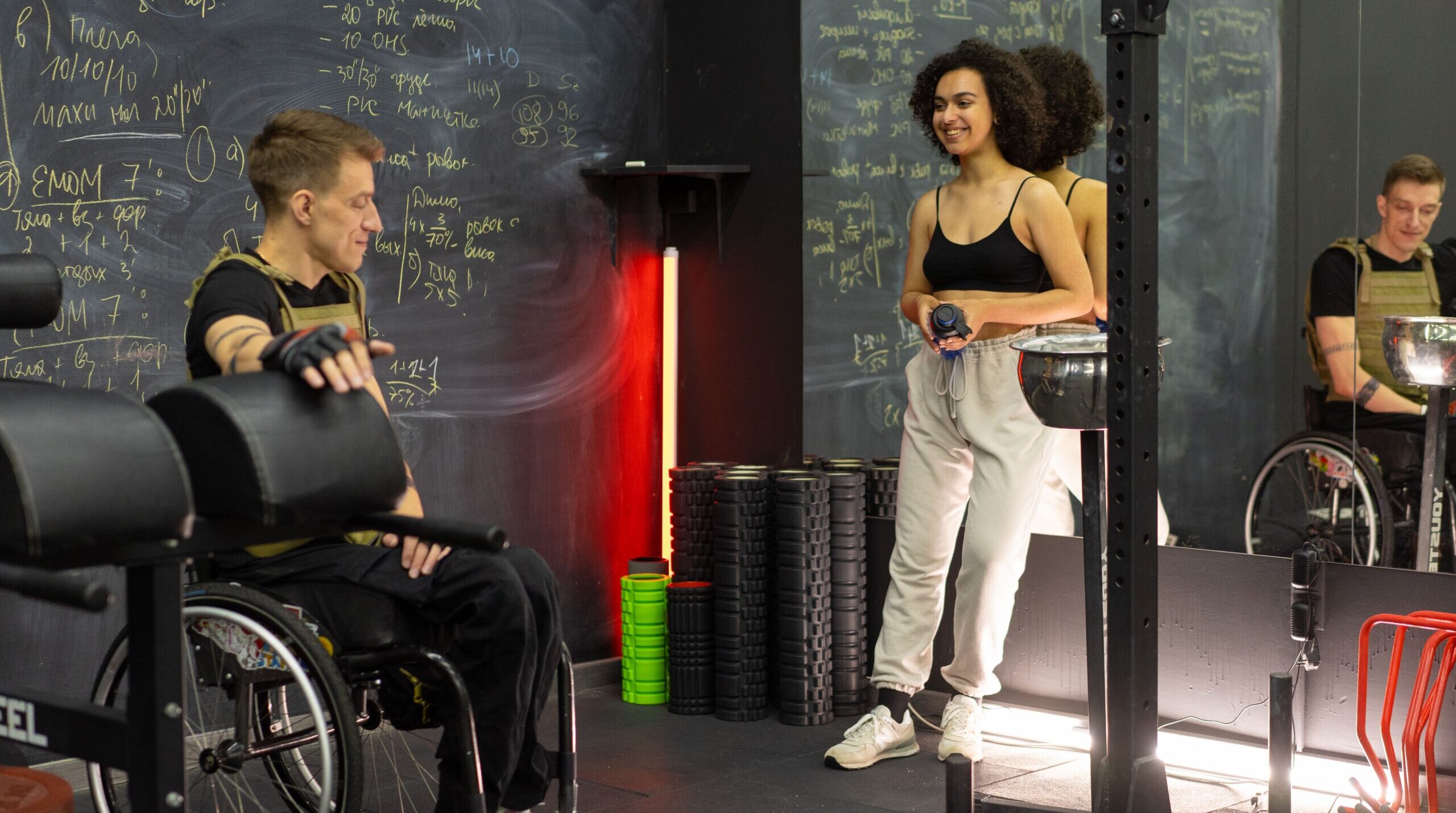 Two people in a gym, one in a wheelchair and one standing, preparing for a workout.